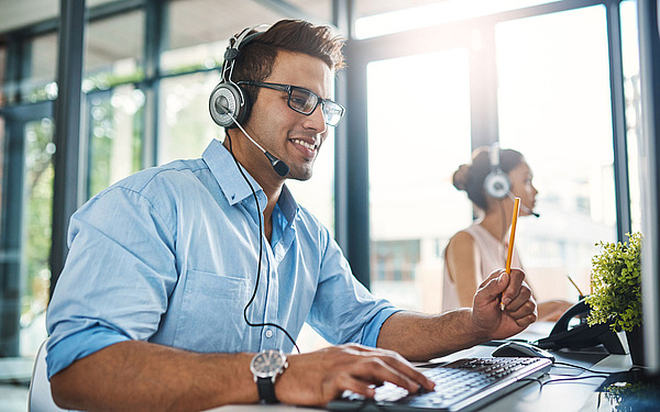 Ein Mann mit Brille und Headset lächelt, während er in einem hellen Büro am Computer arbeitet. Im Hintergrund sitzt eine Frau mit Headset und arbeitet ebenfalls an einem Schreibtisch.