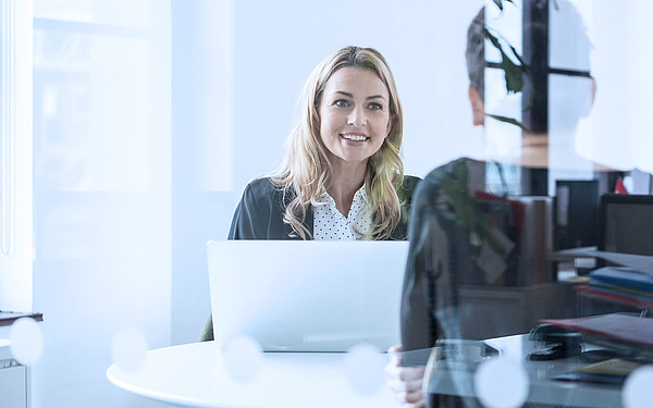 Eine Frau mit blondem Haar sitzt lächelnd mit einem Laptop an einem Schreibtisch in einem Büro. Ihr Gesicht ist einer Person auf der anderen Seite des Schreibtischs zugewandt. Das Büro hat große Fenster und sichtbare Spiegelungen auf der Glaswand.