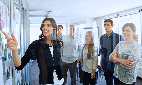 Eine Frau zeigt auf eine Tafel in einem hellen Büro und hält eine Präsentation vor fünf Kollegen, die aufmerksam um sie herum versammelt sind. Bei den Kollegen handelt es sich um eine Mischung aus Männern und Frauen in Berufskleidung, die vor einem großen Fenster stehen.
