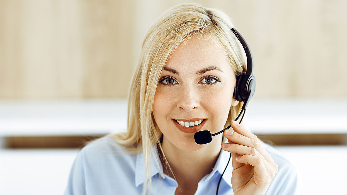 Eine Frau mit blonden Haaren und hellblauem Hemd lächelt und hält ein Headset-Mikrofon. Sie scheint eine Kundendienstmitarbeiterin zu sein. Der Hintergrund ist leicht verschwommen.