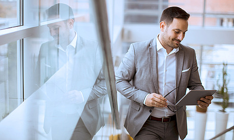 Ein Mann im grauen Anzug hält ein Tablet und eine Brille in der Hand und geht eine helle, moderne Treppe hinauf. Ein großes Fenster spiegelt sein Bild. Er ist auf das Tablet konzentriert und lächelt leicht, was darauf hindeutet, dass er sich mit dem Inhalt beschäftigt.