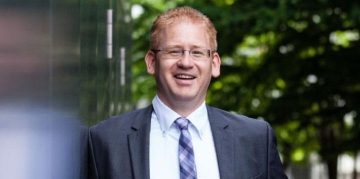 Ein Mann mit kurzen roten Haaren und Brille, der einen grauen Anzug, ein weißes Hemd und eine gemusterte Krawatte trägt, steht lächelnd im Freien, im Hintergrund ist verschwommenes Grün zu sehen.